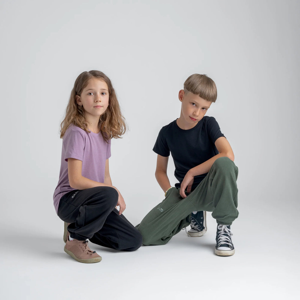 Amo tricou din lână merinos pentru copii; auro-kid-s-fleece-pants-459836-size-banner-large-v-2*: Doi copii în tricou și pantaloni din fleece stau spate în spate, unul dintre ei cu o placă de skateboard