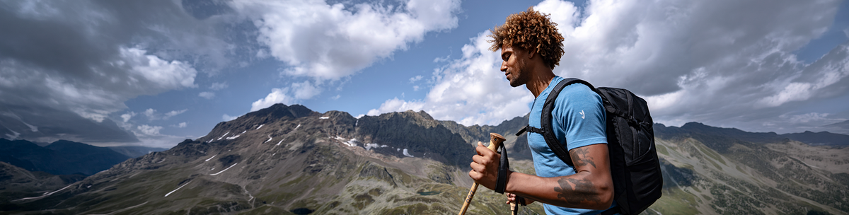 Drumeț în tricou albastru din lână merinos, cu rucsac și băț de trekking în fața peisajului montan