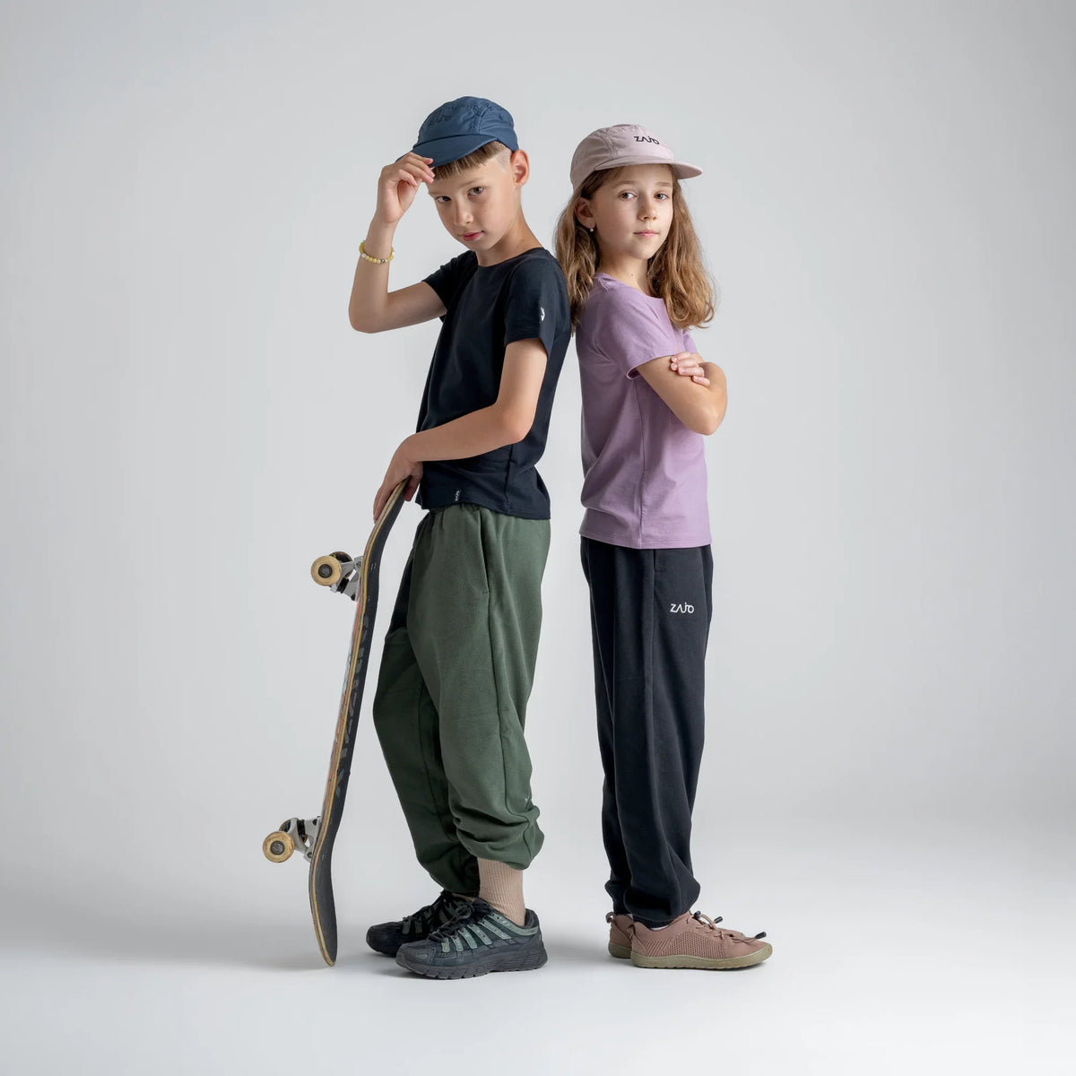 Amo tricou din lână merinos pentru copii; auro-kid-s-fleece-pants-459836-size-banner-large-v-2*: Doi copii în tricou și pantaloni din fleece stau spate în spate, unul dintre ei cu o placă de skateboard